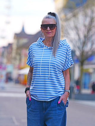 Frau trägt OFF#DLY Ringel m. Kapuze white/lightblue, ein gestreiftes, lässiges Kurzarmshirt mit Kapuze und Tunnelzug. Perfekt für entspannte, sportliche Outfits. Made in Italy.
