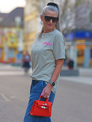 Frau mit Sonnenbrille trägt das OFF#DLY Tee Ibiza taupe, ein lässiges T-Shirt mit IBIZA-Frontdruck. Sie hält eine rote Tasche in der Hand.