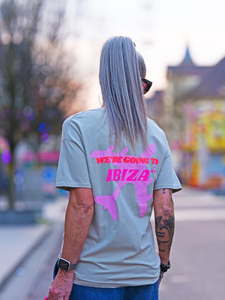 Person mit langen Haaren trägt das OFF#DLY Tee Ibiza taupe, ein graues T-Shirt mit pinkem IBIZA-Schriftzug, im Outdoor-Setting. Perfekt für Sommerlooks, lässig und stilvoll.