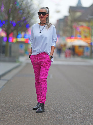 Eine Frau trägt die OFF#DLY Hose Celeste pink mit asymmetrischem Knopfverschluss und dekorativen Reißverschlüssen, gepaart mit schwarzen Stiefeln auf der Straße.
