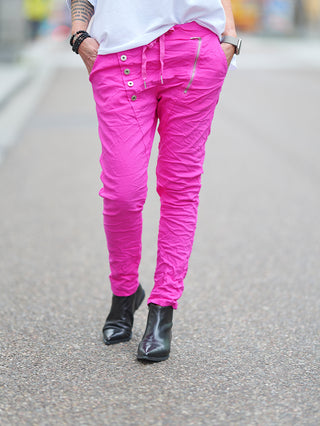 Person trägt die OFF#DLY Hose Celeste pink mit asymmetrischem Knopfverschluss und dekorativen Reißverschlüssen, kombiniert mit schwarzen Stiefeln und einem grauen Armband, auf der Straße.
