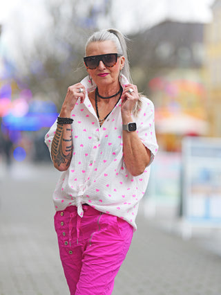 Frau in Sonnenbrille und weißer OFF#DLY Bluse Musselin Heart mit pinken Herzen, Knoten-Detail vorne, lockerer Schnitt, ideal für warme Tage oder Layering, gefertigt aus Baumwolle.