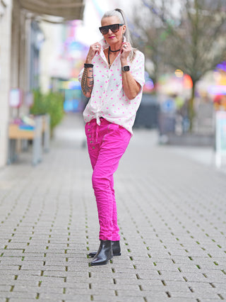 OFF#DLY Bluse Musselin Heart white pink: Frau trägt weiße Bluse mit pinkem Herzmuster und geknoteter Front, kombiniert mit pinker Hose, Sonnenbrille und Stiefeln. Perfekt für lässige, sommerliche Looks.