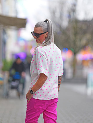 Frau in OFF#DLY Bluse Musselin Heart white pink mit Herzmuster, geknoteter Front und lockerer Passform, kombiniert mit pinker Hose, trägt Sonnenbrille.