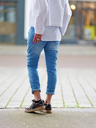 Person trägt OFF#DLY Pant Indria lightblue, eine Denim-Jogpant mit Gürtel und Mini-Tasche, die bequem sitzt. Sichtbar sind blaue Jeans und weißes Oberteil.