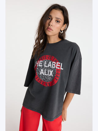 Frau trägt das ALIX The Label Tee charcoal, ein oversized T-Shirt mit auffälligem Print auf der Vorderseite, kombiniert mit roten Hosen und langen Haaren. Perfekt für lässigen Streetstyle.