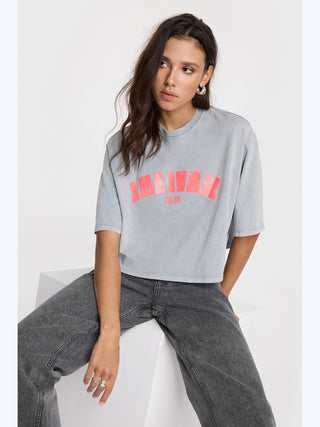 Frau sitzt auf einem weißen Würfel, trägt ein ALIX The Label Tee in Hellgrau. Das Shirt hat roten Text und besteht aus 100% Baumwolle, perfekt für einen lässigen Look.