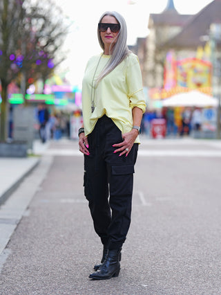 Frau trägt OFF#DLY Lightsweat Calista gelb, einen leichten Boxy-Sweater mit Used-Look, gelben Rundhalsausschnitt, kombiniert mit schwarzen Hosen und schwarzen Stiefeln auf dem Bürgersteig.