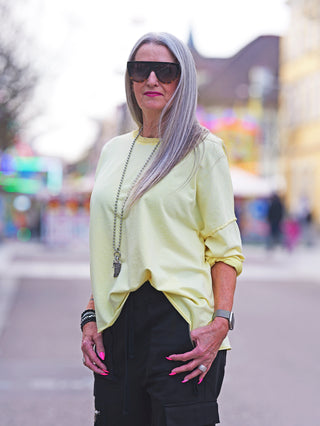 Frau mit grauem Haar trägt den OFF#DLY Lightsweat Calista gelb, einen legeren Boxy-Sweater mit Used-Look, sichtbar an Kragen und Nähten, kombiniert mit einer Sonnenbrille.
