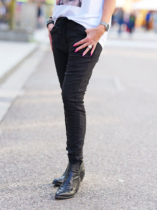 Produktbild der OFF#DLY Pant Ava black, zeigt schwarze Crinkle-Hose mit subtilen Paisley-Stickereien, kombiniert mit schwarzen Stiefeletten. Die Hose hat einen elastischen Bund und Taschen.