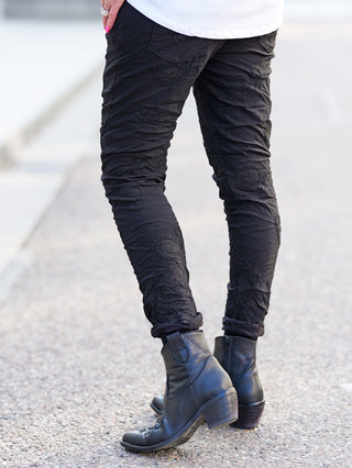 OFF#DLY Pant Ava black: Schwarze Crinkle-Hose mit subtiler Paisley-Stickerei und elastischem Bund, getragen mit schwarzen Stiefeln. Ideal für lässige, stilvolle Auftritte.