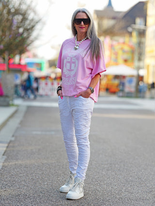 Frau trägt das OFF#DLY Sweater Milano rose, ein lässiges Oversize-Shirt mit „Old School Milano“-Print, kombiniert mit weißen Hosen und Sneakers auf dem Gehweg.
