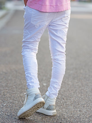 OFF#DLY Pant Ava white: Weiße bestickte Crinkle-Hose getragen von einer Person, kombiniert mit weißen Schuhen. Die Hose zeigt feine Paisley-Stickereien und bietet durch den elastischen Bund Komfort.