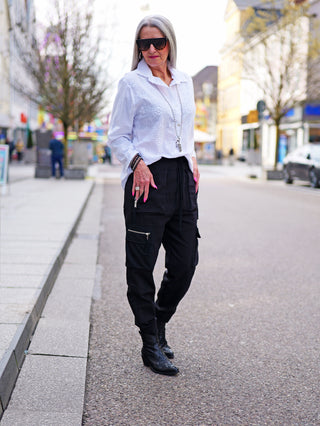 Frau trägt die OFF#DLY Bluse Tiana weiß mit floraler Lochstickerei, Oversize-Passform, kombiniert mit schwarzer Hose und Stiefeln, während sie auf dem Bürgersteig steht.