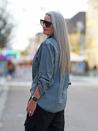 Frau mit langen grauen Haaren trägt die OFF#DLY Bluse Tiana anthrazit, bestickt mit Blumenmustern, kombiniert mit Sonnenbrille und Armbanduhr, verkörpert italienische Eleganz und Boho-Charme.