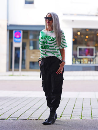Frau trägt OFF#DLY Ringelsweat Monaco white/green mit grünen und weißen Streifen, 3D-Schriftzug „MONACO 64 FRANCE“. Kombiniert mit schwarzen Hosen und Stiefeln auf einem Gehweg.
