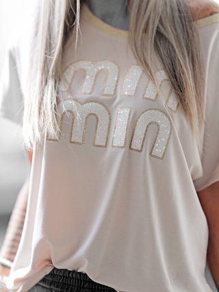 Nahaufnahme des OFF#DLY Shirt Min Min beige, ein elegantes Damenoberteil mit glitzerndem Rundhalsbündchen und strukturiertem min min Schriftzug, ideal für stilvolle Anlässe.