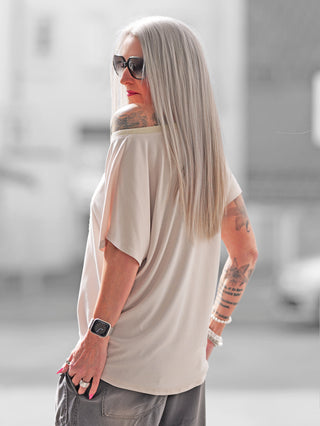 Frau mit langen Haaren trägt OFF#DLY Shirt Min Min beige, zeigt Glitzerdetails und strukturierten min min-Schriftzug, kombiniert mit Sonnenbrille und Tattoos, ideal für Alltag und Anlässe.