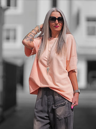 Frau trägt den OFF#DLY Sweat Lola apricot, ein oversize Kurzarmsweater mit Raw-Edge-Saum, kombiniert mit Sonnenbrille und grauer Hose, ideal für lässige, modische Sommerlooks.