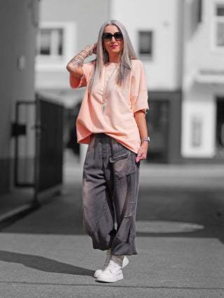 Frau trägt den OFF#DLY Sweat Lola apricot, einen oversized Kurzarmsweater mit Raw-Edge-Saum, kombiniert mit weißen Stiefeln, ideal für einen lässigen, modernen Look.