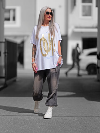 Frau trägt ein OFF#DLY Long Tee OUI in Weiß/Gold, während sie auf dem Bürgersteig geht. Das Shirt hat goldene Buchstaben als Designmerkmal und wird mit grauen Hosen kombiniert.