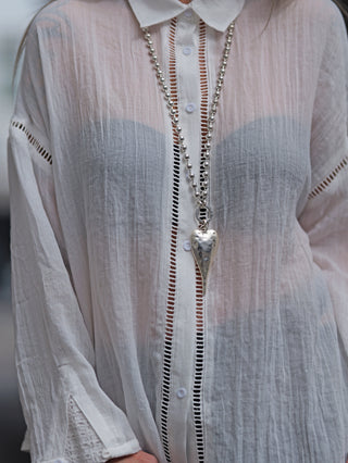 Frau trägt die OFF#DLY Bluse Viola white, eine weiße Oversize-Bluse mit Hohlsaum-Details, Dekorative Knopfleiste und entspannter Silhouette, kombiniert mit einem silbernen Herzanhänger.