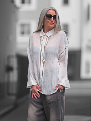 Frau trägt OFF#DLY Bluse Viola white, eine weiße Oversize-Bluse mit Hohlsaum-Details, kombiniert mit Sonnenbrille, betont durch dekorative Einsätze an Ärmeln und Knopfleiste.