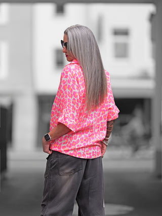 Frau mit grauem Haar trägt die OFF#DLY Bluse Musselin Liora Leo pink, eine luftige Musselinbluse mit Leopardenmuster und Knopfleiste, ideal für sommerliche Anlässe.