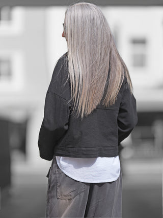 OFF#DLY Jacke Tiana schwarz, oversized Denim-Style, boxy geschnitten, sichtbar von hinten an einer Frau getragen, mit langen Haaren. Druckknöpfe und Brusttaschen betonen den Look. Made in Italy.