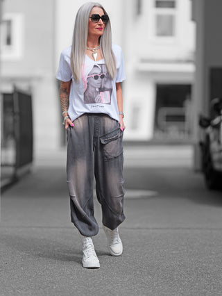 Frau trägt OFF#DLY Tee Valencia weiß/pink mit I don't care-Slogan, tiefem V-Ausschnitt. Kombiniert mit Sonnenbrille und grauer Hose auf der Straße, lässig und stilvoll.