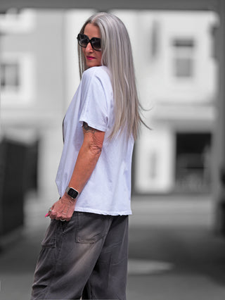 Frau trägt OFF#DLY Tee Valencia weiß/beige mit I don't care-Slogan, V-Ausschnitt. Sonnenbrille und Tattoo sichtbar. Perfekt für lässigen Look.