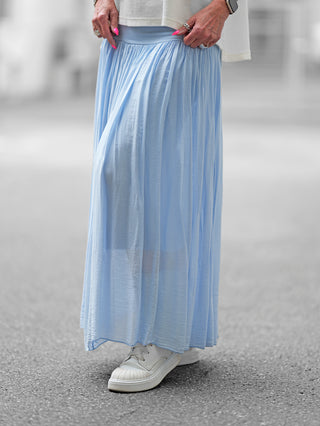 Person trägt den OFF#DLY Rock Sienna lightblue, ein fließender Maxirock mit weitem Bund und leicht gekräuseltem Stoff, kombiniert mit weißen Schuhen. Ideal für stilvolles Auftreten.