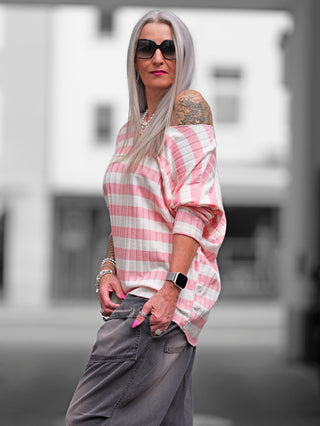Frau in einem OFF#DLY Ringel Strick beige/rose, mit gestreiftem Muster und weicher Rippstruktur. Sie trägt das Shirt lässig, kombiniert mit modischen Accessoires wie Sonnenbrille und Armbanduhr.