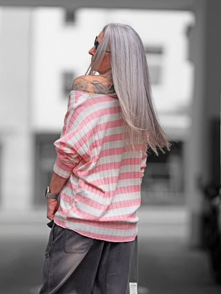 Frau mit grauen Haaren trägt ein OFF#DLY Ringel Strick beige/rose, ein gestreiftes Shirt mit Rippstruktur und femininen Schnitt, kombiniert mit grauen Hosen und Tattoos auf der Schulter.