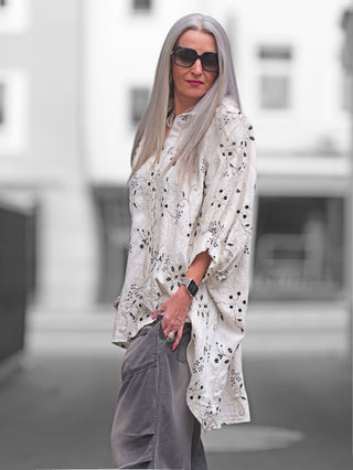 Frau trägt die OFF#DLY Longbluse Stick beige black mit Oversize-Schnitt und Stickereien, kombiniert mit Sonnenbrille, zeigt stilvollen Look und Bewegungsfreiheit.