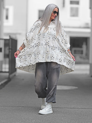Frau trägt eine OFF#DLY Longbluse Stick beige black mit kunstvollen Stickereien, weitem Oversize-Schnitt und klassischem Hemdblusenkragen, kombiniert mit grauen Hosen und weißen Stiefeln.