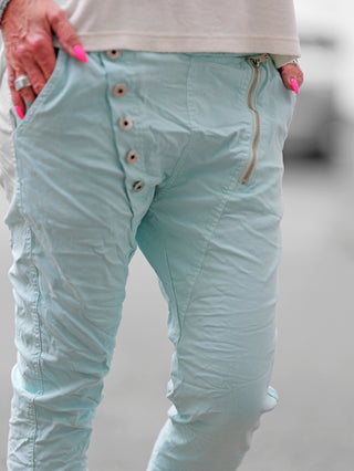 Die OFF#DLY Hose Celeste türkis ist eine weiße Crinkle-Hose mit asymmetrischem Knopfverschluss und dekorativen Reißverschlüssen, ideal für einen modernen, vielseitigen Look bis Größe 42.