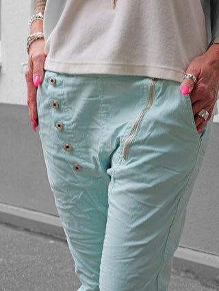 Die OFF#DLY Hose Celeste türkis zeigt einen leichten Crinkle-Effekt mit asymmetrischem Knopfverschluss und dekorativen Reißverschlüssen, ideal für einen modernen, lässigen Look.