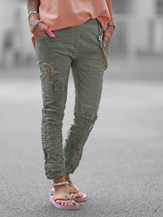 OFF#DLY Pant Vesper khaki: Eine Person trägt graue Jogpants mit Paillettenpatches und dekorativen Kordel-Chain-Elementen. Die Hose hat einen elastischen Bund und Hände in den Taschen.