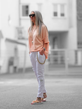 Frau trägt OFF#DLY Pant Vesper white mit Paillettenpatches, lässigem Schnitt, elastischem Bund und Kordel-Chain-Elementen, kombiniert mit Sonnenbrille und pfirsichfarbenem Shirt.