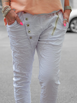 Person trägt OFF#DLY Hose Celeste weiß mit asymmetrischem Knopfverschluss und dekorativen Reißverschlüssen. Crinkle-Effekt verleiht modernen Look. Stretchiges Material bietet Komfort und Passform.