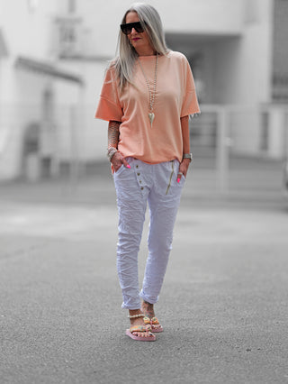 Frau trägt OFF#DLY Hose Celeste weiß mit Crinkle-Effekt und asymmetrischem Knopfverschluss auf der Straße, kombiniert mit weißen Sandalen, für einen lässigen, modernen Look.