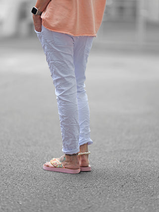 OFF#DLY Hose Celeste weiß: Eine Person trägt die weiße Crinkle-Hose mit asymmetrischem Knopfverschluss und dekorativen Reißverschlüssen, kombiniert mit rosa Sandalen. Ideal für einen modernen, lässigen Look.