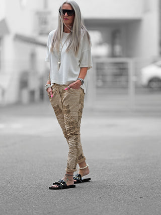 Frau trägt OFF#DLY Print Pant Lindsey camel mit Crinkle-Look und Silk-Touch auf der Straße, kombiniert mit schwarzen Sandalen. Ideal für stilvolle, entspannte City-Looks.