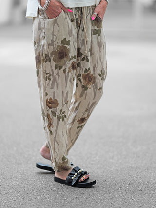 Person trägt OFF#DLY Linen Pant Romy beige/khaki mit floralem Print. Weite Passform, elastischer Bund, seitliche Taschen. Kombiniert mit Sandalen für einen lässigen Sommerlook.