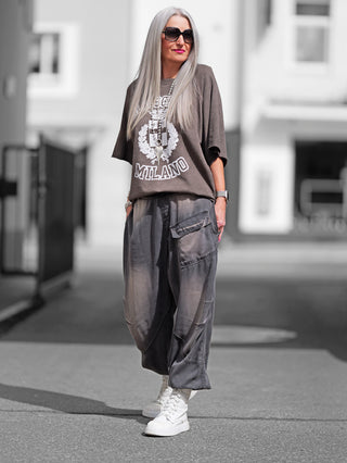 Frau trägt OFF#DLY Sweater Milano olive, ein lässiges Oversize-Shirt mit „Old School Milano“-Print, kombiniert mit grauer Hose und weißen Schuhen, komplettiert durch große Sonnenbrille und Statement-Kette.