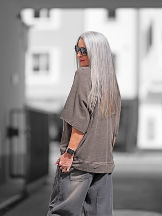 Frau mit langen grauen Haaren trägt das OFF#DLY Sweater Milano olive, ein lässiges Oversize-Shirt mit Old School Milano-Print, kombiniert mit Sonnenbrille und Uhr.