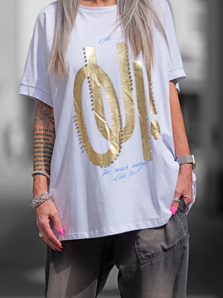 Frau trägt das OFF#DLY Long Tee OUI in Weiß/Gold, mit goldenen Buchstaben auf der Vorderseite. Der lässige Schnitt und das modische Design machen es ideal für den Alltag.