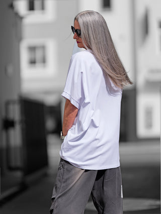 Eine Frau mit langen grauen Haaren und Sonnenbrille trägt das OFF#DLY Long Tee OUI in Weiß/Gold. Sie steht draußen und zeigt den lässigen Stil des Shirts.