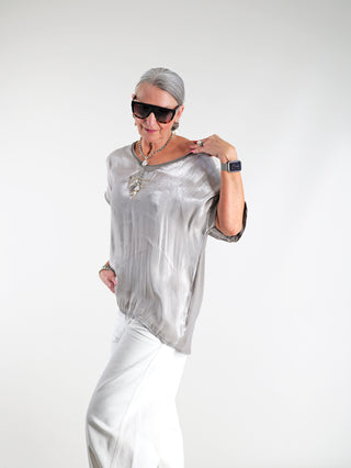Frau trägt das OFF#DLY Tee Wake up taupe mit silbernem Glanz vorne und goldfarbenem Statement-Print. Sie kombiniert es mit Sonnenbrille und Kette, betont den lässig-eleganten Look.